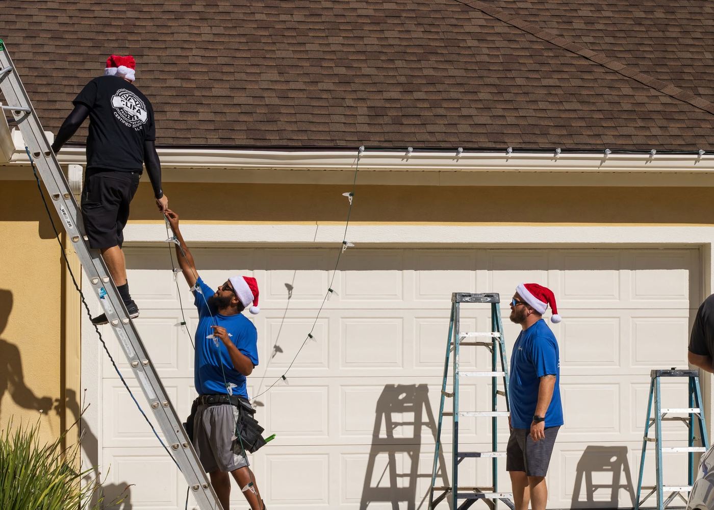 Three Five Star crew members in Santa hats hanging lights along a garage roofline